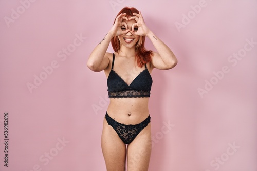 Young caucasian woman wearing lingerie over pink background doing ok gesture like binoculars sticking tongue out, eyes looking through fingers. crazy expression. © Krakenimages.com