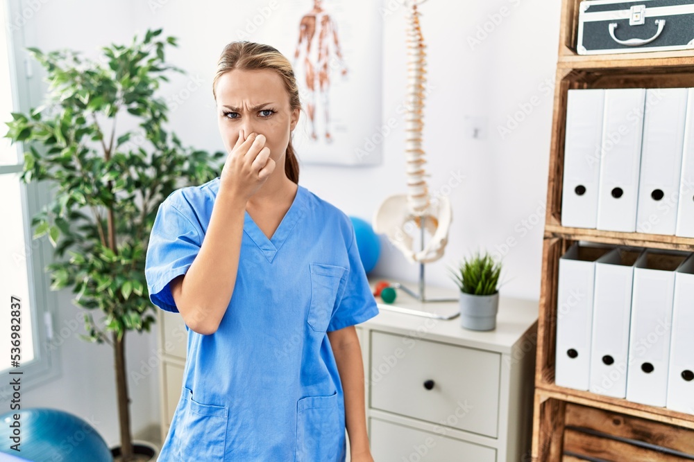 Young caucasian woman working at pain recovery clinic smelling something stinky and disgusting, intolerable smell, holding breath with fingers on nose. bad smell Young caucasian woman working at pain recovery clinic smelling something stinky and disgusting, intolerable smell, holding breath with fingers on nose. bad smell