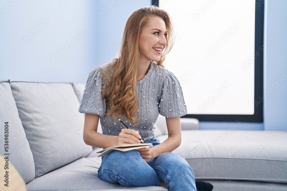 Young blonde woman writing on notebook sitting on sofa at home Young blonde woman writing on notebook sitting on sofa at home