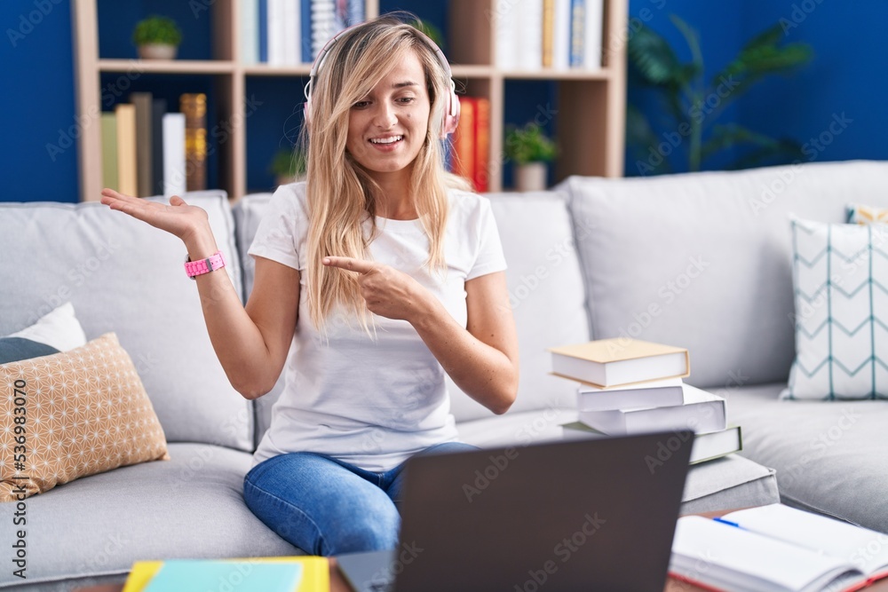 Young blonde woman studying using computer laptop at home amazed and smiling to the camera while presenting with hand and pointing with finger. Young blonde woman studying using computer laptop at home amazed and smiling to the camera while presenting with hand and pointing with finger.