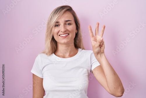 Young blonde woman standing over pink background showing and pointing up with fingers number three while smiling confident and happy.