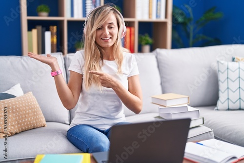 Young blonde woman studying using computer laptop at home amazed and smiling to the camera while presenting with hand and pointing with finger.