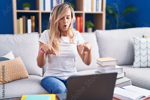Young blonde woman studying using computer laptop at home pointing down with fingers showing advertisement, surprised face and open mouth