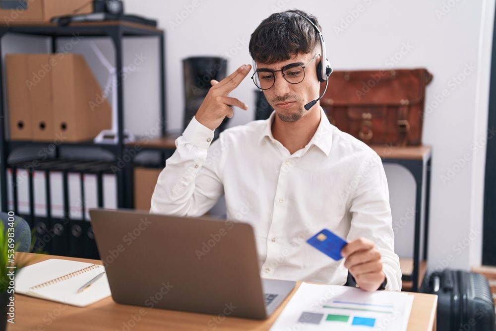 Young hispanic man working using computer laptop holding credit card shooting and killing oneself pointing hand and fingers to head like gun, suicide gesture. Young hispanic man working using computer laptop holding credit card shooting and killing oneself pointing hand and fingers to head like gun, suicide gesture.