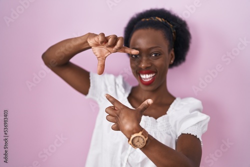 African woman with curly hair standing over pink background smiling making frame with hands and fingers with happy face. creativity and photography concept.