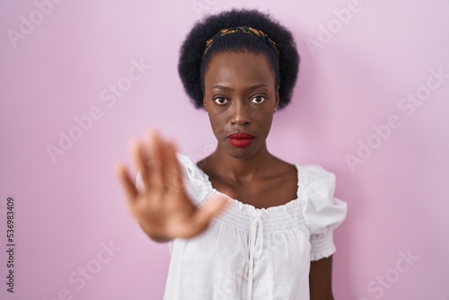 African woman with curly hair standing over pink background doing stop sing with palm of the hand. warning expression with negative and serious gesture on the face.