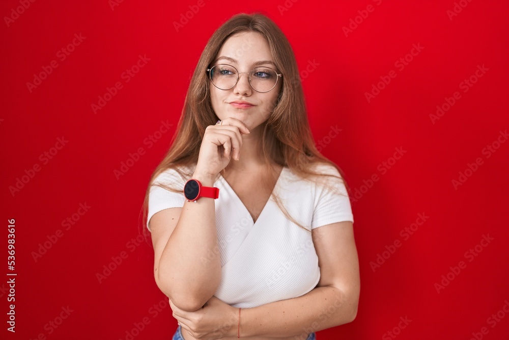 Young caucasian woman standing over red background with hand on chin thinking about question, pensive expression. smiling and thoughtful face. doubt concept. Young caucasian woman standing over red background with hand on chin thinking about question, pensive expression. smiling and thoughtful face. doubt concept.