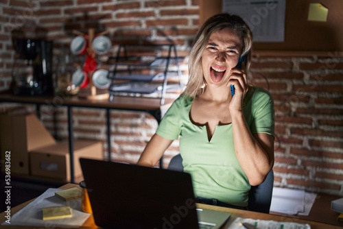 Young beautiful woman working at the office at night speaking on the phone sticking tongue out happy with funny expression. emotion concept.