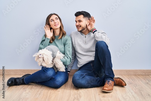 Young hispanic couple sitting on the floor with dog smiling with hand over ear listening an hearing to rumor or gossip. deafness concept.