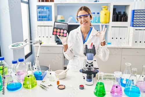 Young blonde woman working at scientist laboratory with make up doing ok sign with fingers, smiling friendly gesturing excellent symbol