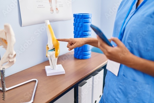 Young hispanic woman physiotherapist using smartphone pointing to anatomical model of knee at rehab clinic