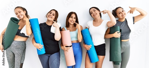 Group of women holding yoga mat standing over isolated background smiling cheerful showing and pointing with fingers teeth and mouth. dental health concept.