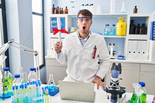 Young hispanic man working at scientist laboratory holding diploma scared and amazed with open mouth for surprise  disbelief face