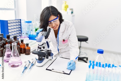 Young latin woman wearing scientist uniform using microscope write on clipboard at laboratory