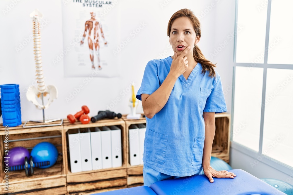Middle age hispanic physiotherapist woman working at pain recovery clinic looking fascinated with disbelief, surprise and amazed expression with hands on chin Middle age hispanic physiotherapist woman working at pain recovery clinic looking fascinated with disbelief, surprise and amazed expression with hands on chin