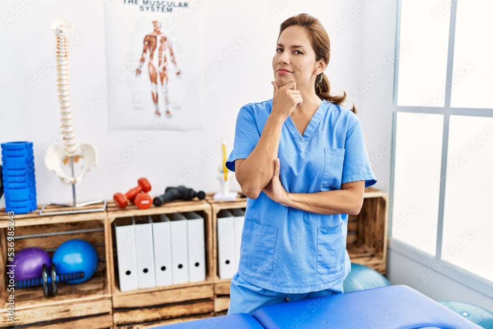 Middle age hispanic physiotherapist woman working at pain recovery clinic looking confident at the camera smiling with crossed arms and hand raised on chin. thinking positive. Middle age hispanic physiotherapist woman working at pain recovery clinic looking confident at the camera smiling with crossed arms and hand raised on chin. thinking positive.