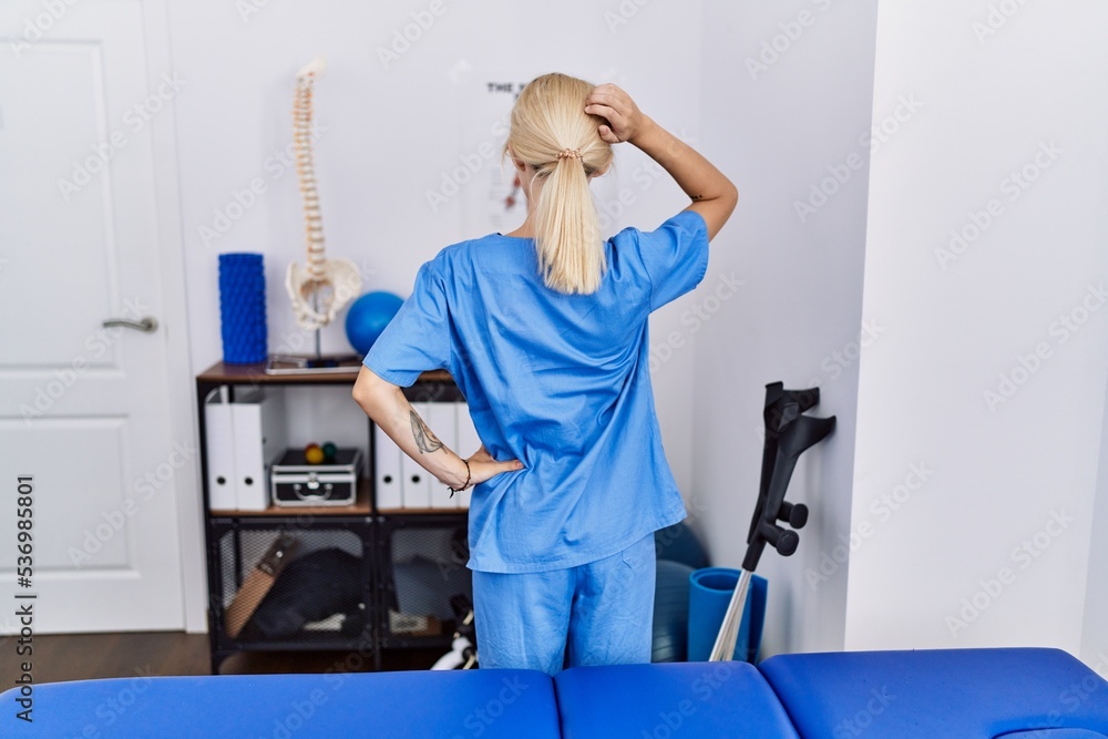 Young physiotherapist woman working at pain recovery clinic backwards thinking about doubt with hand on head