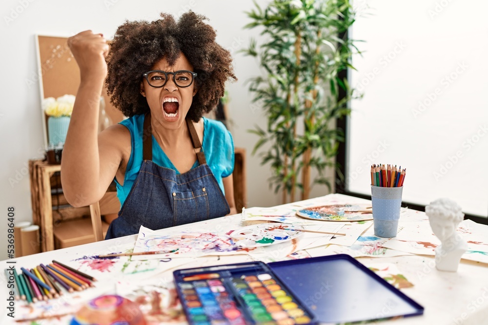Fototapeta premium Beautiful african american woman with afro hair painting at art studio angry and mad raising fist frustrated and furious while shouting with anger. rage and aggressive concept.