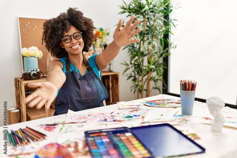 Fototapeta premium Beautiful african american woman with afro hair painting at art studio looking at the camera smiling with open arms for hug. cheerful expression embracing happiness.