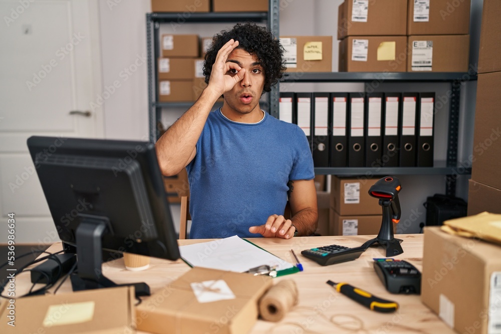 Hispanic man with curly hair working at small business ecommerce doing ok gesture shocked with surprised face, eye looking through fingers. unbelieving expression. Hispanic man with curly hair working at small business ecommerce doing ok gesture shocked with surprised face, eye looking through fingers. unbelieving expression.