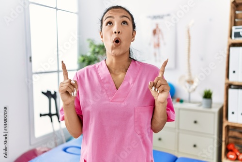 Young hispanic woman working at pain recovery clinic amazed and surprised looking up and pointing with fingers and raised arms.