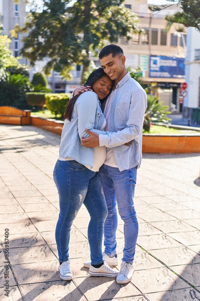 Fototapeta premium Young latin couple expecting baby hugging each other standing at park