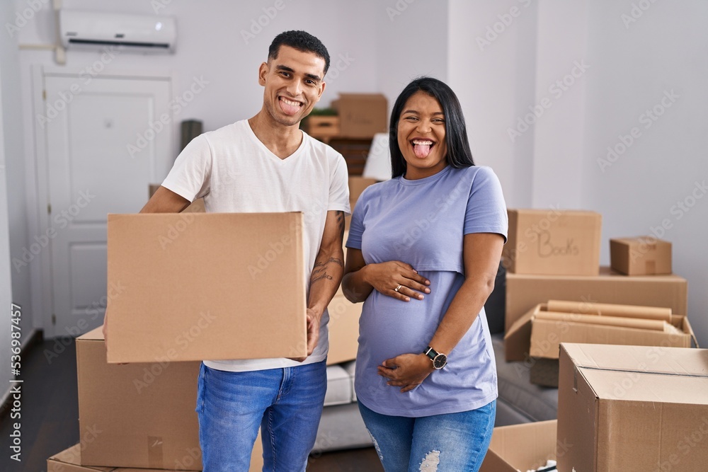 Young hispanic couple expecting a baby moving to a new home sticking tongue out happy with funny expression. Young hispanic couple expecting a baby moving to a new home sticking tongue out happy with funny expression.