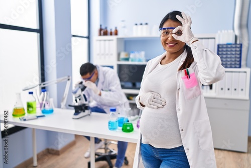 Young hispanic woman expecting a baby working at scientist laboratory smiling happy doing ok sign with hand on eye looking through fingers