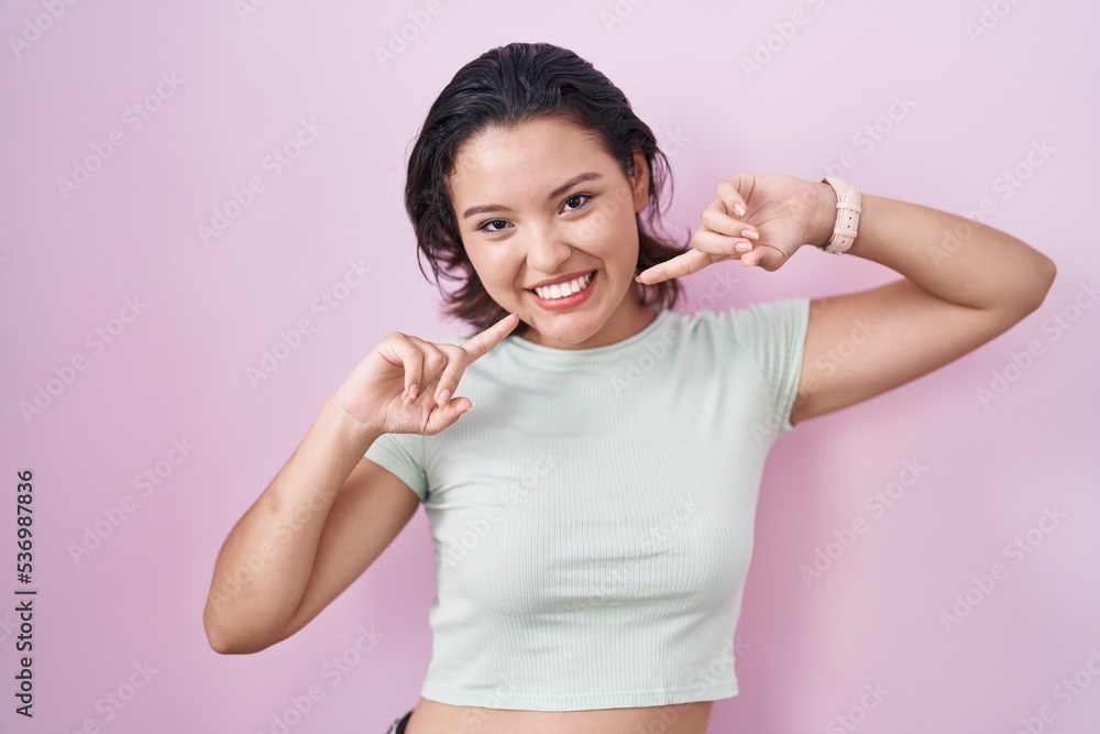 Hispanic young woman standing over pink background smiling cheerful showing and pointing with fingers teeth and mouth. dental health concept. Hispanic young woman standing over pink background smiling cheerful showing and pointing with fingers teeth and mouth. dental health concept.