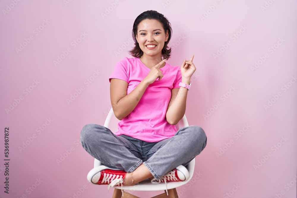 Hispanic young woman sitting on chair over pink background smiling and looking at the camera pointing with two hands and fingers to the side. Hispanic young woman sitting on chair over pink background smiling and looking at the camera pointing with two hands and fingers to the side.