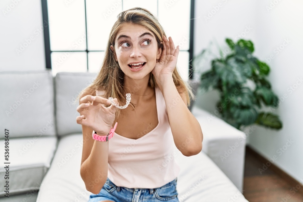 Fototapeta premium Young blonde woman holding invisible aligner orthodontic smiling with hand over ear listening an hearing to rumor or gossip. deafness concept.