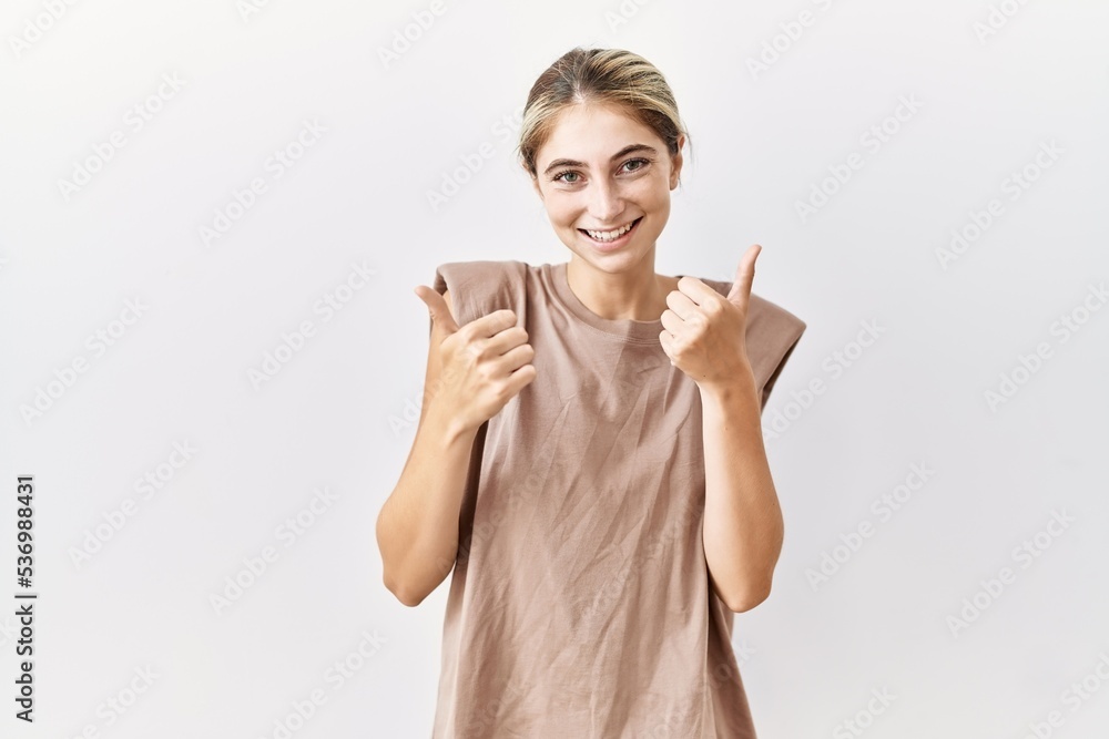 Young blonde woman standing over isolated background success sign doing positive gesture with hand, thumbs up smiling and happy. cheerful expression and winner gesture. Young blonde woman standing over isolated background success sign doing positive gesture with hand, thumbs up smiling and happy. cheerful expression and winner gesture.