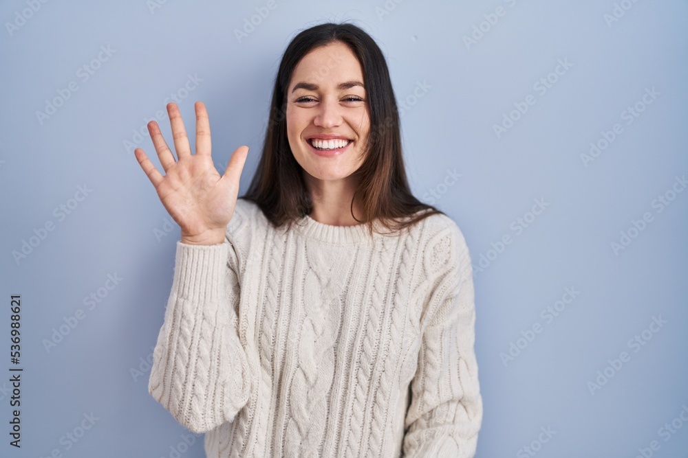 Young brunette woman standing over blue background showing and pointing up with fingers number five while smiling confident and happy. Young brunette woman standing over blue background showing and pointing up with fingers number five while smiling confident and happy.