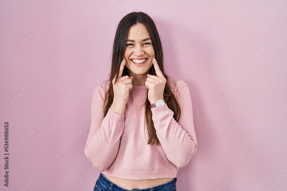 Young brunette woman standing over pink background smiling with open mouth, fingers pointing and forcing cheerful smile Young brunette woman standing over pink background smiling with open mouth, fingers pointing and forcing cheerful smile