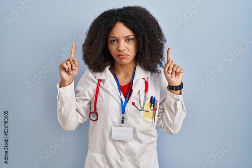 Young african american woman wearing doctor uniform and stethoscope pointing up looking sad and upset  indicating direction with fingers  unhappy and depressed.