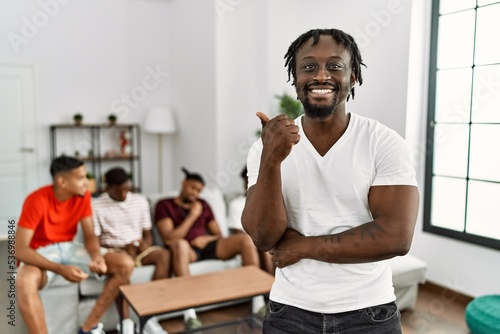 Young african man with friends at the living room smiling with happy face looking and pointing to the side with thumb up.