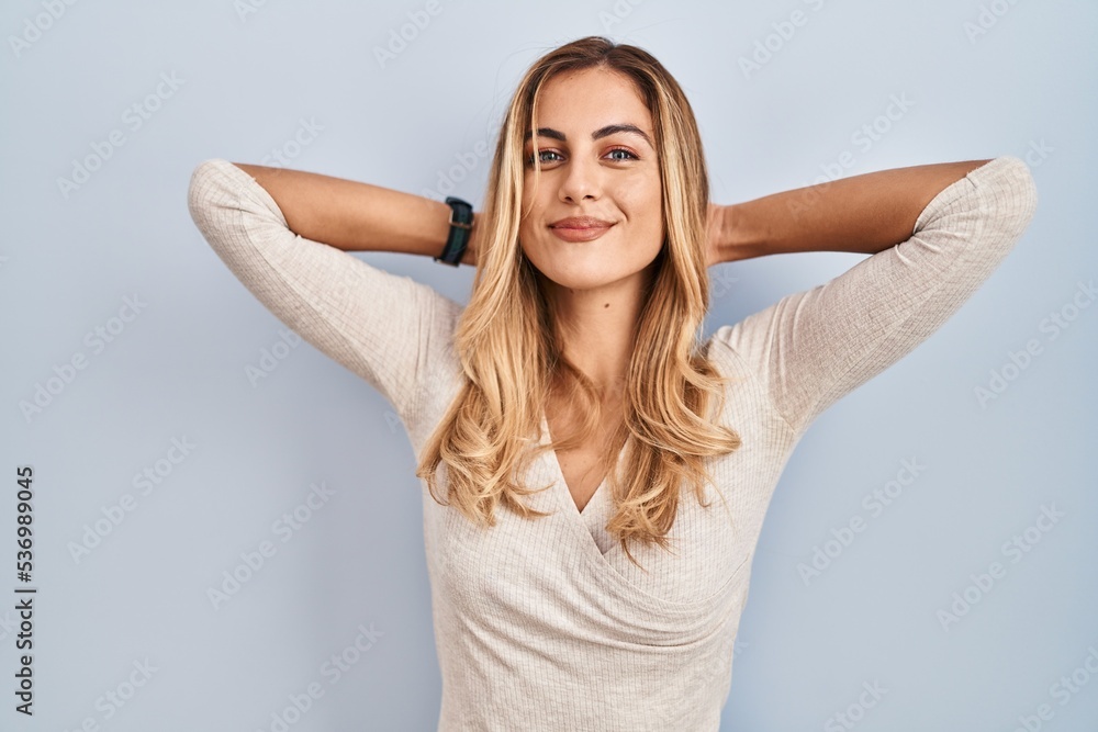 Young blonde woman standing over isolated background relaxing and stretching, arms and hands behind head and neck smiling happy