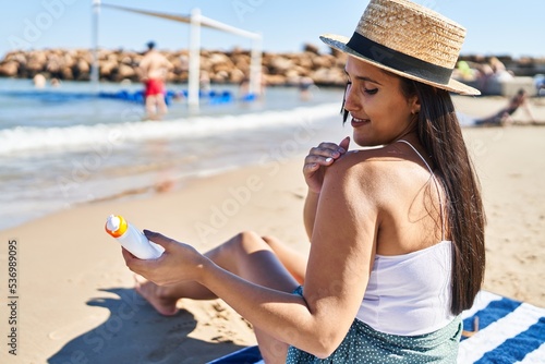 Young hispanic woman using sunscreen sitting on sand at seaside