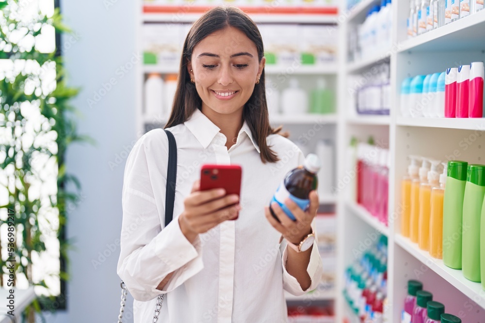 Young beautiful hispanic woman customer using smartphone holding medicine bottle at pharmacy