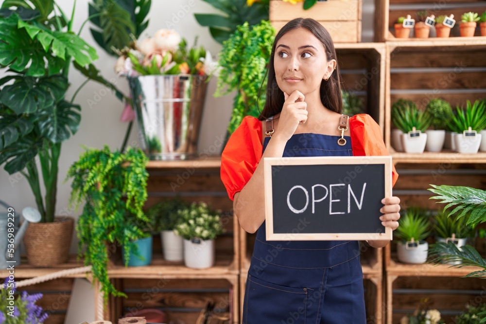 Young hispanic woman working at florist holding open sign serious face thinking about question with hand on chin, thoughtful about confusing idea Young hispanic woman working at florist holding open sign serious face thinking about question with hand on chin, thoughtful about confusing idea