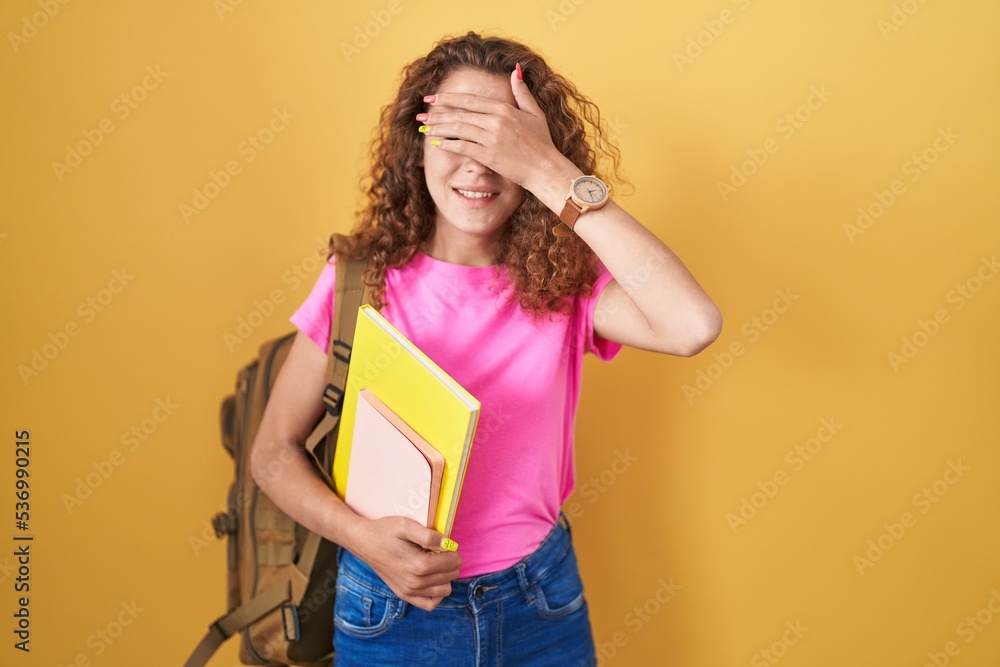 Young caucasian woman wearing student backpack and holding books smiling and laughing with hand on face covering eyes for surprise. blind concept. Young caucasian woman wearing student backpack and holding books smiling and laughing with hand on face covering eyes for surprise. blind concept.