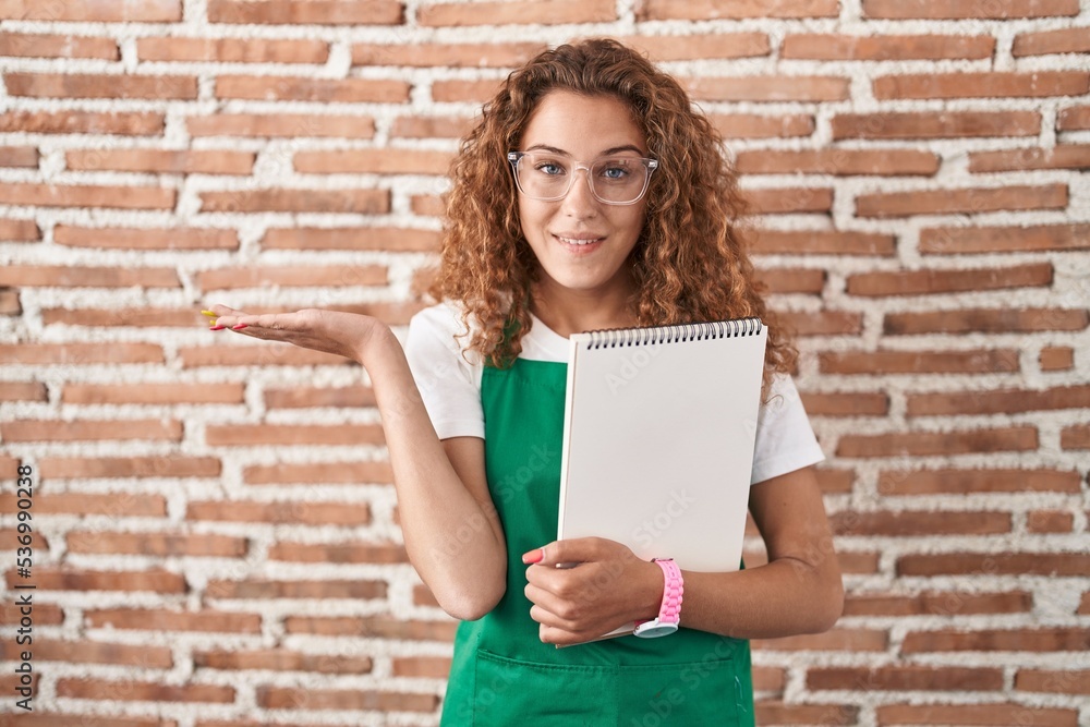 Young caucasian woman holding art notebook smiling cheerful presenting and pointing with palm of hand looking at the camera. Young caucasian woman holding art notebook smiling cheerful presenting and pointing with palm of hand looking at the camera.