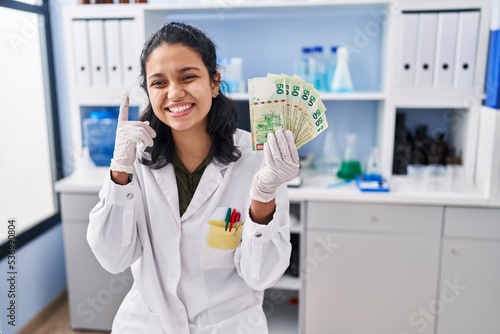 Hispanic woman with dark hair working at scientist laboratory holding money surprised with an idea or question pointing finger with happy face  number one