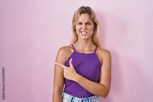 Young blonde woman standing over pink background pointing aside worried and nervous with forefinger  concerned and surprised expression