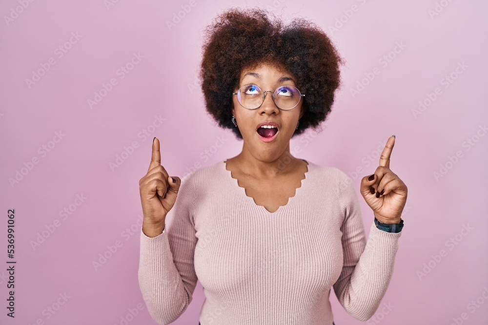 Young african american woman standing over pink background amazed and surprised looking up and pointing with fingers and raised arms. Young african american woman standing over pink background amazed and surprised looking up and pointing with fingers and raised arms.