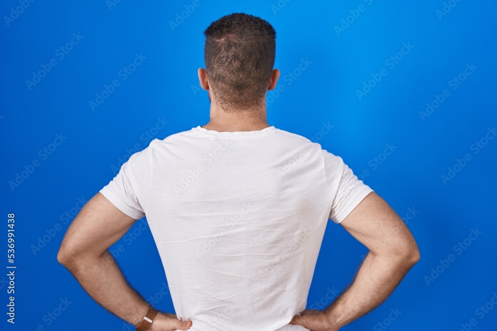 Young caucasian man standing over blue background standing backwards looking away with arms on body Young caucasian man standing over blue background standing backwards looking away with arms on body