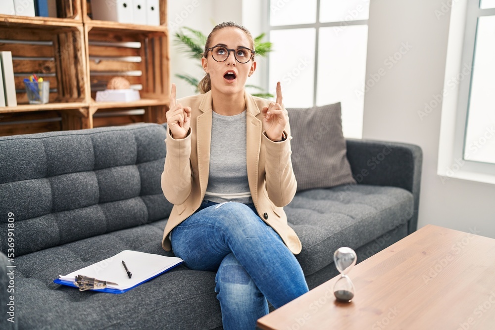 Young woman working at consultation office amazed and surprised looking up and pointing with fingers and raised arms. Young woman working at consultation office amazed and surprised looking up and pointing with fingers and raised arms.