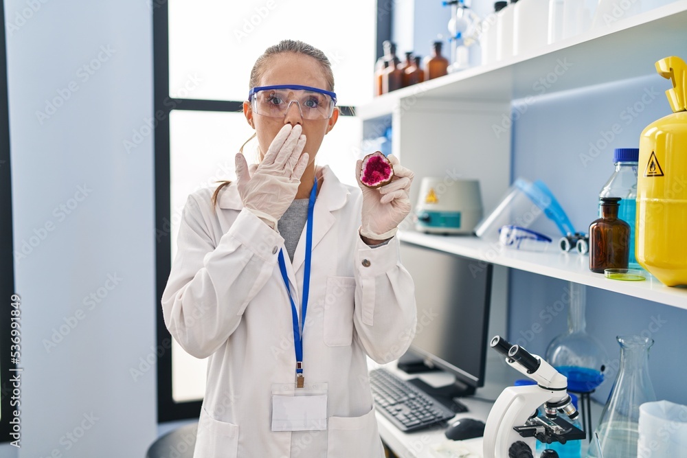 Young woman working at scientist laboratory holding geode covering mouth with hand, shocked and afraid for mistake. surprised expression Young woman working at scientist laboratory holding geode covering mouth with hand, shocked and afraid for mistake. surprised expression