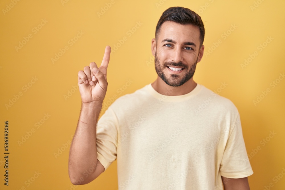 Handsome hispanic man standing over yellow background showing and pointing up with finger number one while smiling confident and happy. Handsome hispanic man standing over yellow background showing and pointing up with finger number one while smiling confident and happy.