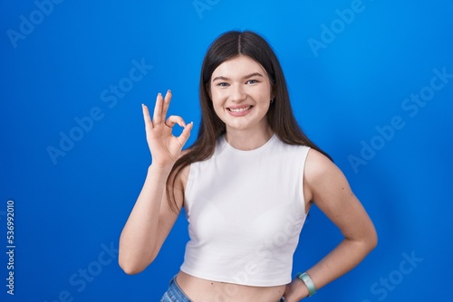 Young caucasian woman standing over blue background smiling positive doing ok sign with hand and fingers. successful expression.